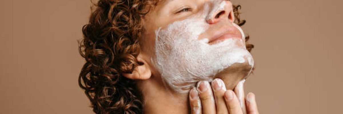 Handsome man enjoying beauty treatment while applying face cream and standing on brown background